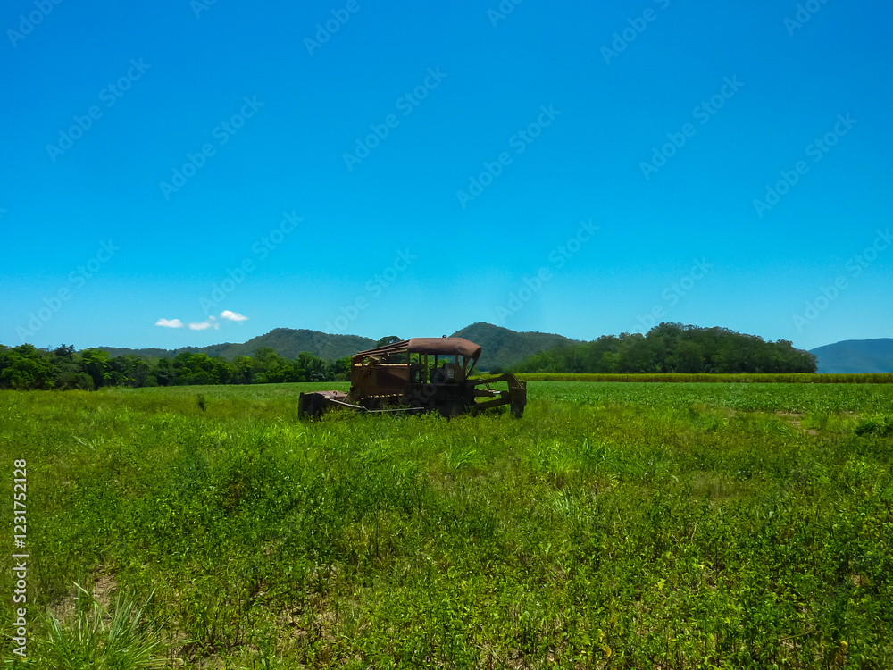 Old abandoned, piece of farm machinery sits in green field in Cairns, Queensland, Australia. Machinery, which may be a tractor or harvester, is surrounded by tall grass. Hills in remote landscape Old abandoned, piece of farm machinery sits in green field in Cairns, Queensland, Australia. Machinery, which may be a tractor or harvester, is surrounded by tall grass. Hills in remote landscape