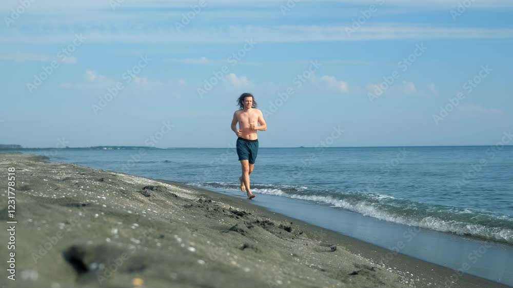 Long-haired athletic man running barefoot on sea sandy beach on vacations. Moving with freedom enjoying ocean waves beneath bright sunny sky. Sport outdoors activity healthy lifestyle. Travel tourism. Long-haired athletic man running barefoot on sea sandy beach on vacations. Moving with freedom enjoying ocean waves beneath bright sunny sky. Sport outdoors activity healthy lifestyle. Travel tourism.