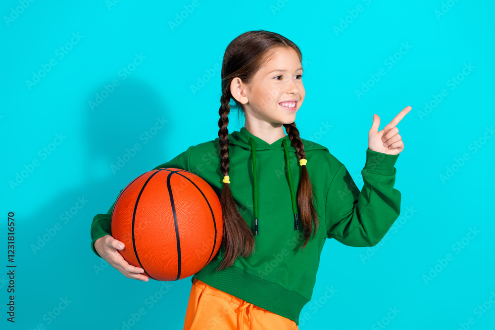 Smiling girl in green hoodie holding basketball and pointing joyfully against turquoise background Smiling girl in green hoodie holding basketball and pointing joyfully against turquoise background