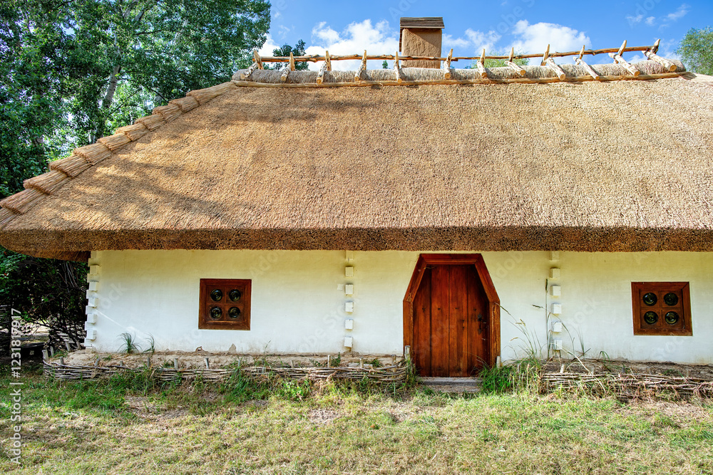 Rustic thatched roof cottage in the old Ukrainian style nestled in a tranquil forest clearing under a bright blue sky.