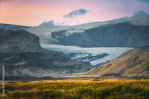 Vue panoramique sur un glacier islandais descendant des montagnes sous une lumière dorée. Contraste entre la glace, la roche volcanique et la végétation, capturant la majesté brute de l’Islande. photo