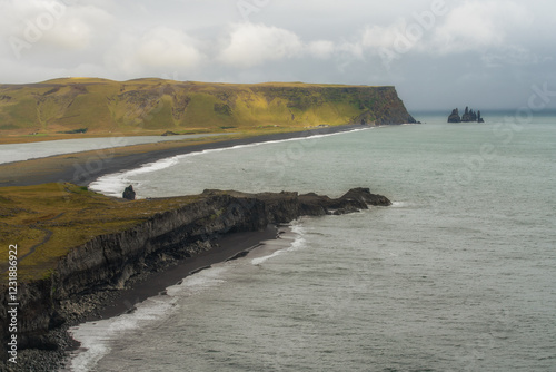Vue panoramique sur la plage de sable noir de Reynisfjara en Islande, avec ses falaises abruptes et les formations rocheuses de Reynisdrangar émergeant de l’Atlantique sous une lumière dramatique.