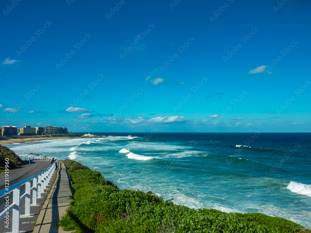 Fototapeta premium Scenic coastal walkway along Newcastle Beach, NSW, Australia, featuring turquoise ocean waves, white railings, and lush green vegetation. Modern beachfront buildings line the horizon