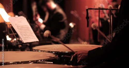 A close-up view of a timpani player with drumsticks poised over the instrument, and sheet music visible on a stand. The blurred background captures other orchestra musicians