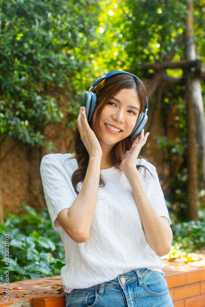 Attractive young Asian woman sits in a park, wearing wireless headphones and smiling, listening to music while relaxing in a tranquil park. Joyful Asian woman feeling relaxed, happy, and stress-free. Attractive young Asian woman sits in a park, wearing wireless headphones and smiling, listening to music while relaxing in a tranquil park. Joyful Asian woman feeling relaxed, happy, and stress-free.
