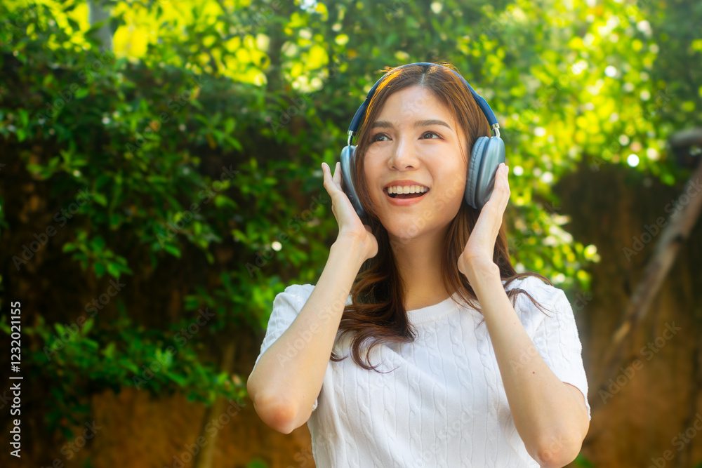 Attractive young Asian woman sits in a park, wearing wireless headphones and smiling, listening to music while relaxing in a tranquil park. Joyful Asian woman feeling relaxed, happy, and stress-free. Attractive young Asian woman sits in a park, wearing wireless headphones and smiling, listening to music while relaxing in a tranquil park. Joyful Asian woman feeling relaxed, happy, and stress-free.