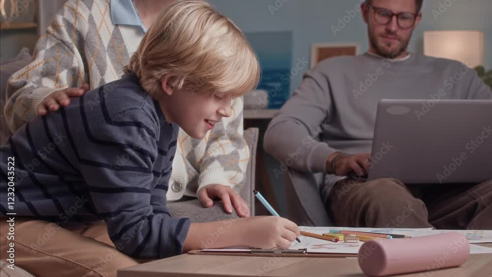 Handheld shot of Caucasian mom and her son doing art homework at table while father working on wireless laptop in background at home