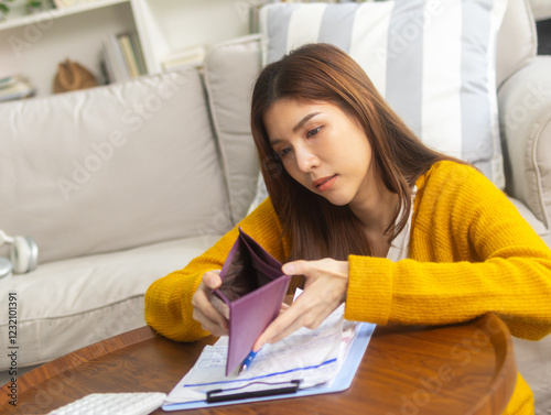 Frustrated young Asian woman looks at her empty wallet with a worried expression, feeling stressed about her financial instability and unpaid loan. Serious Asian woman thinking about financial future.