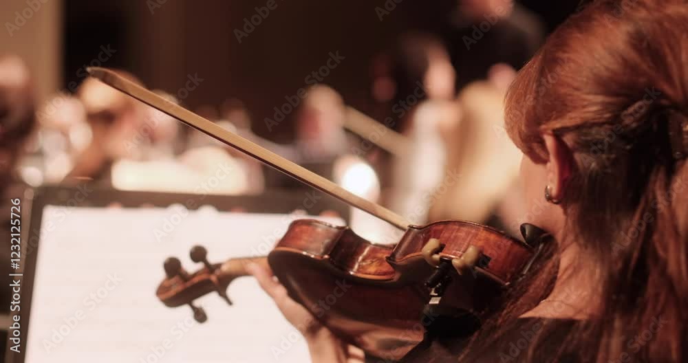 A close-up view of a violin player during a rehearsal of an academic classical orchestra in a softly lit concert hall. The conductor and other musicians are visible in the background
