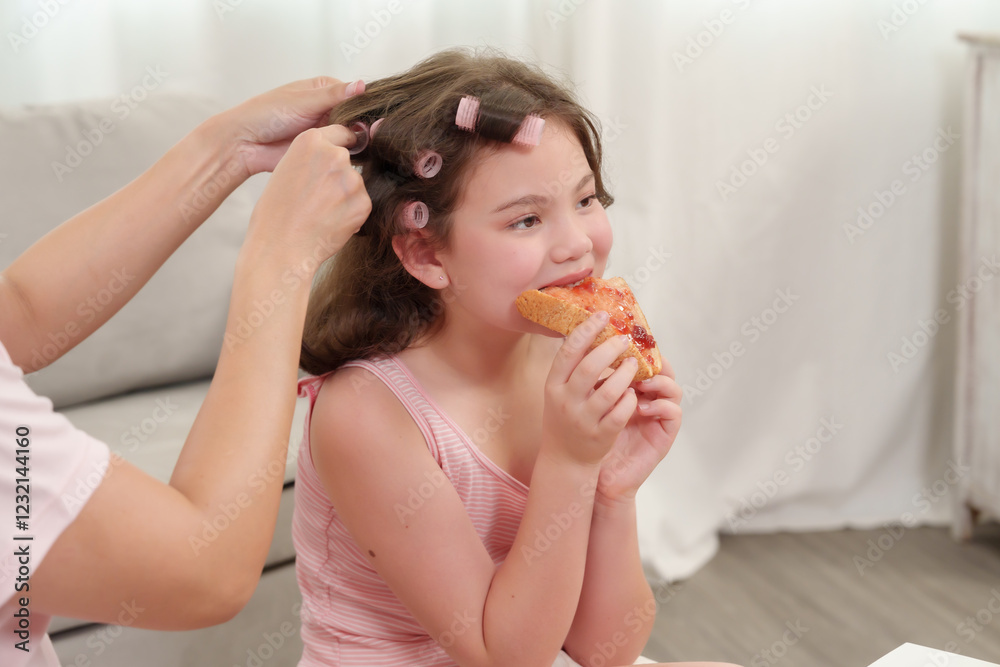 Happiness of Caucasian daughter and Asian mother. Mother curling daughter's hair while she enjoys a pastry. Smiling girl appreciating beauty time and snack. Warm family moment at home. Happiness of Caucasian daughter and Asian mother. Mother curling daughter's hair while she enjoys a pastry. Smiling girl appreciating beauty time and snack. Warm family moment at home.