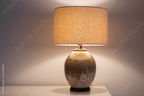 Selective focus view of lighted vintage ceramic table lamp with floral hand-painted motif set on corner of pale table against plain wall photo