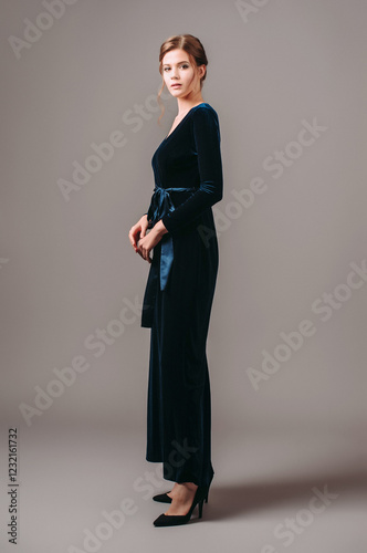 Wrap maxi velour evening dress in navy blue. Long sleeve velvet gown with bow knot. Studio portrait of elegant young woman on high heels, fashionable outfit for an event. Pretty brunette lady