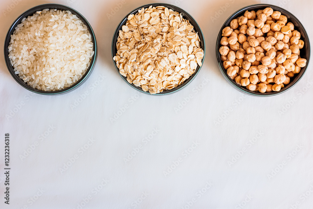 Three ceramic bowls filled with oats, rice, and chickpeas on a white surface, with scattered grains and legumes creating a natural, rustic food composition Three ceramic bowls filled with oats, rice, and chickpeas on a white surface, with scattered grains and legumes creating a natural, rustic food composition