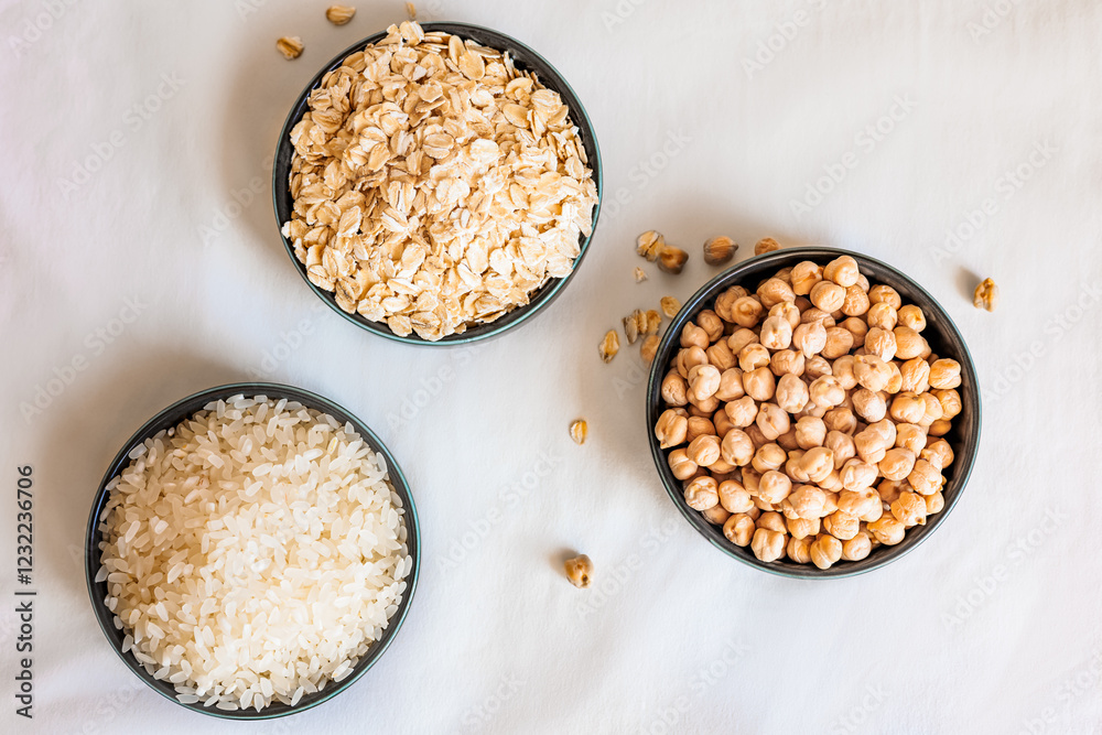 Top view of three ceramic bowls with oats, rice, and chickpeas arranged in a triangular composition on a soft white background, with scattered grains adding a natural touch Top view of three ceramic bowls with oats, rice, and chickpeas arranged in a triangular composition on a soft white background, with scattered grains adding a natural touch