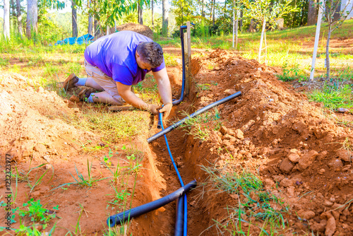 Worker is installing black irrigation PVC water pipes in freshly dug trench while working outdoors.