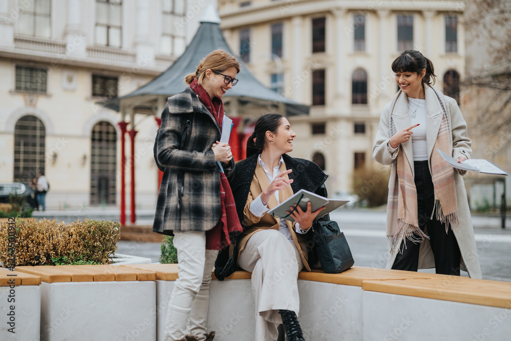 Fototapeta premium Three women with documents discussing an idea outdoors near modern architecture, portraying teamwork and cooperation in a professional setting while sharing a friendly and relaxed moment.
