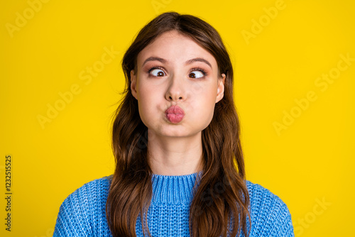Funny young woman puffing cheeks in a blue sweater against a vibrant yellow wall