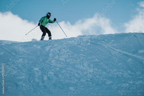 A person is skiing downhill on a snowy slope, surrounded by a clear blue sky and bright sunlight. The skier wears a green jacket, using poles, displaying outdoor adventure and winter sports activity.
