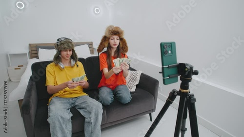 Two young influencers funny fluffy hats with cash throwing in a cozy room while filming content for their social media channel in the afternoon