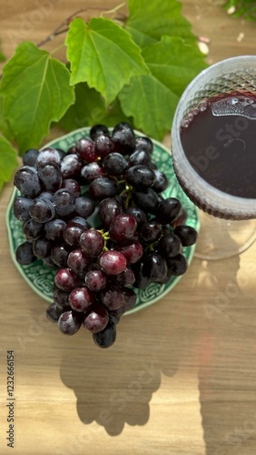 A close-up of fresh, dark purple grapes with water droplets, arranged in a green ceramic cup and plate. Vibrant green leaves add contrast, with a woven basket in the softly blurred background.