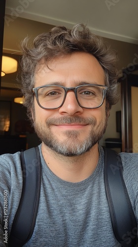 Man with curly hair and glasses smiles indoors, showcasing relaxed atmosphere...