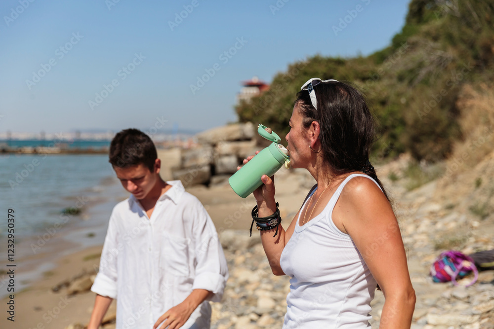 A mother holds a reusable water bottle while her teenage son adjusts her sunglasses, enjoying a lighthearted moment on a rocky seaside under bright sun