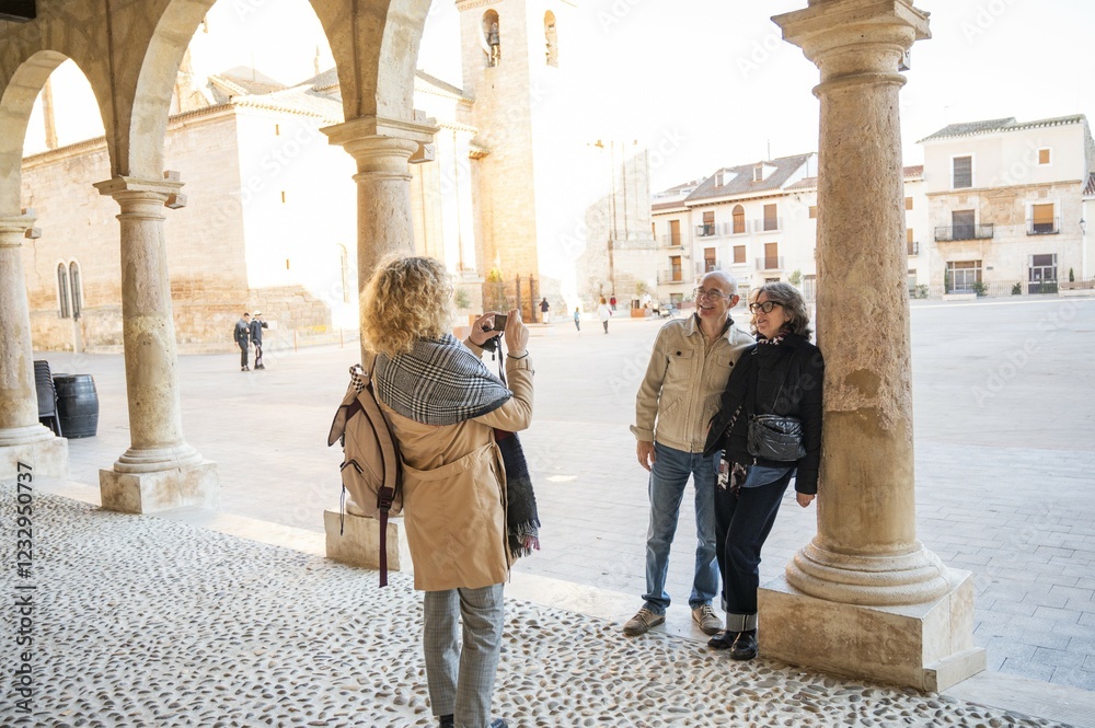 Fototapeta premium Middle-aged woman photographing her friends posing next to stone columns in a historic Spanish plaza during a sunny day