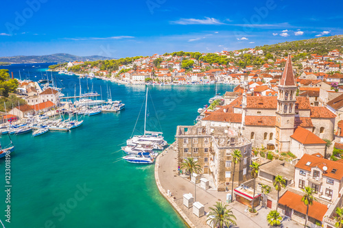 A drone view of Milna on Brač Island, Croatia, featuring the Church of Our Lady of Annunciation and its bell tower, a scenic bay filled with boats, a vibrant waterfront, and traditional Mediterranean 