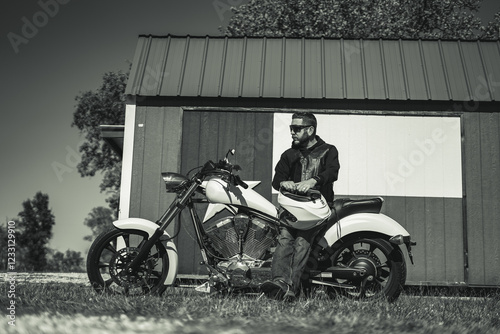 Man In Leather Jacket Sitting on Motorcycle and Holding Helmet In Front of Texas Flag Shed Black and White photo