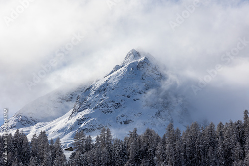 Alpine landscape with snow-capped mountains immersed in clouds and larch forests in Engadine Valley, Canton of Grisons - Switzerland photo