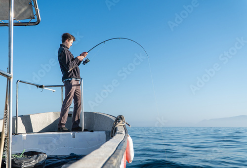 Obraz na płótnie A young man focuses on casting his fishing line from a boat into the serene waters, immersed in the peaceful atmosphere of a calm day at sea. w salonie