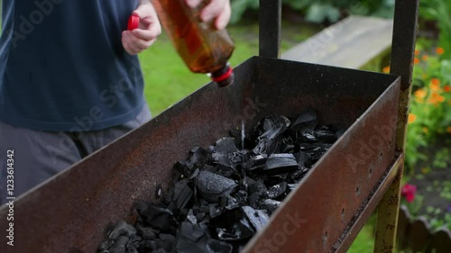 Professional chef preparing outdoor barbecue, carefully pouring lighter fluid onto black coals in metal brazier, readying grill for summer cooking session