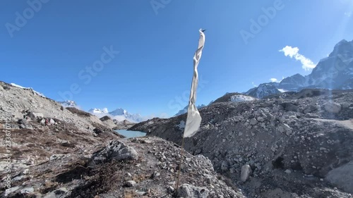 A white prayer flag flutters in the strong wind atop a flagpole planted in the vast Ngozumba Glacier, a symbol of spirituality and resilience amidst the harsh beauty of the Himalayas