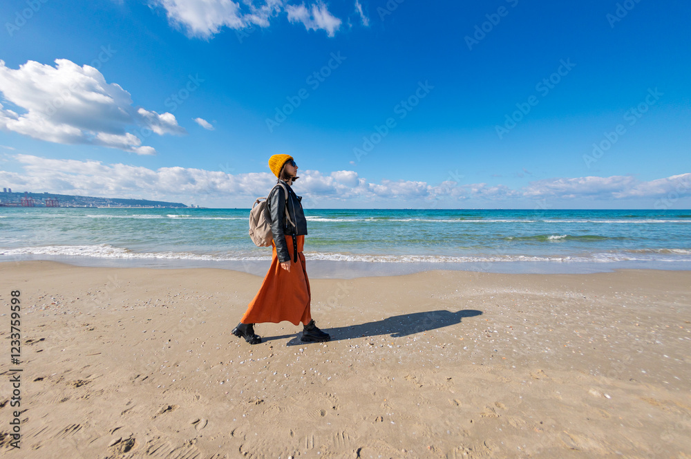 Traveling woman with backpack on the beach in cold weather. Stylish woman enjoying a walk near the sea on a sunny day. Traveling woman with backpack on the beach in cold weather. Stylish woman enjoying a walk near the sea on a sunny day.