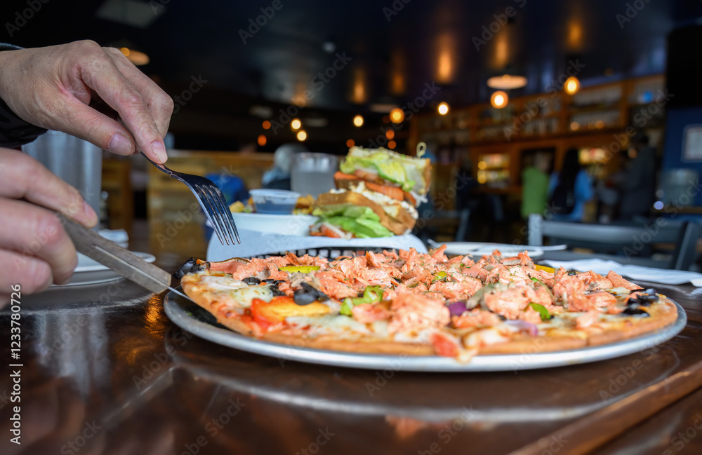 Hands holding forks and cutting a salmon pizza. Unrecognizable people in the restaurant. Alaska.