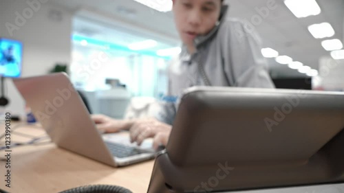A focused office worker engages in a phone conversation while managing tasks on a laptop. The modern, open workspace reflects a collaborative business environment.