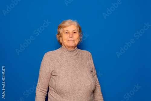 Horizontal portrait of a Caucasian woman in her 70s posing proudly and satisfied with the stage of her life she is living, isolated on a blue studio background.