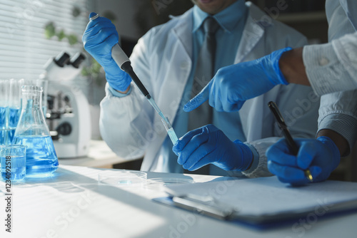Professional chemists wearing lab coats and gloves are conducting experiment using pipette and test tube, taking notes and discussing results in modern laboratory