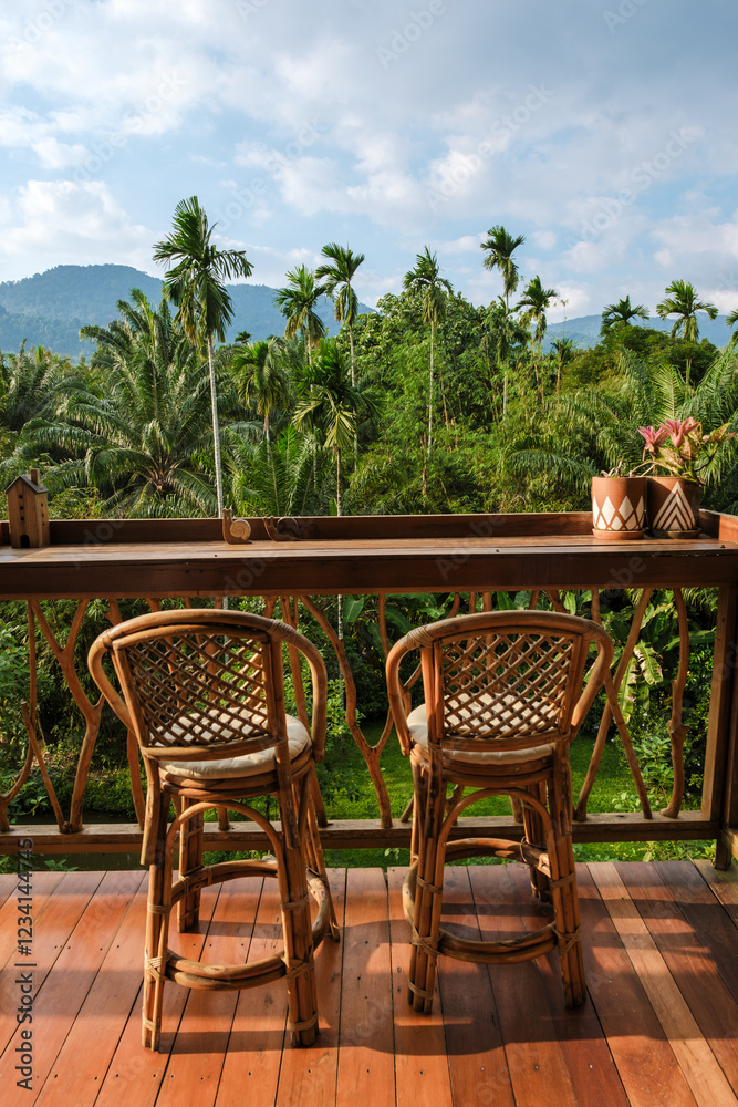Obraz premium Two elegant bamboo chairs sit on a wooden deck, overlooking a vibrant tropical landscape filled with palm trees and mountains in the distance. This peaceful spot invites relaxation and tranquility.
