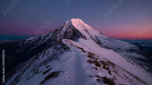 Snow covered mountain peak with rugged terrain, glowing sky at sunset or dawn photo