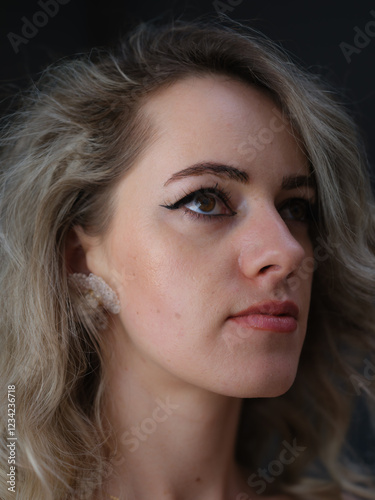 A close-up portrait of a stylish woman with wavy blonde hair, elegant silver rings, and soft makeup, gazing confidently at the camera against a dark background