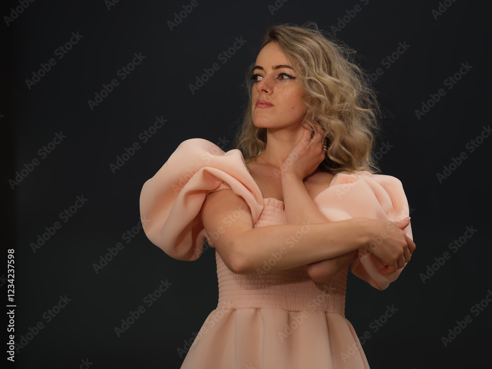 A stylish woman in a pink dress with puffed sleeves poses elegantly with her arms crossed, gazing to the side with confidence and grace against a dark background