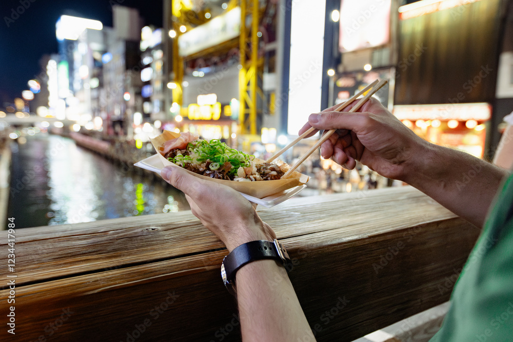 A tourist is taking a takoyaki ball with chopsticks, a popular street food in Osaka, Japan. It's a summer night, the lights of the billboards are reflecting on the canal water in the background