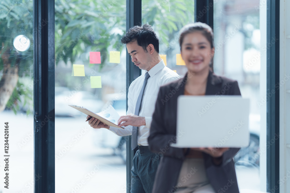 Businesswoman with Laptop in Modern Office: A smiling businesswoman confidently holds a laptop, while a male colleague reviews documents in the background.
