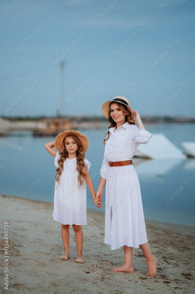 a girl and a young mother in white dresses and hats stand barefoot on the sand on the seashore after sunset a girl and a young mother in white dresses and hats stand barefoot on the sand on the seashore after sunset