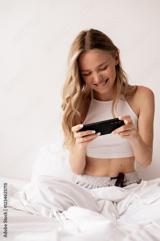 Beautiful caucasian young blonde woman playing game online using smartphone sitting on bed in bedroom, hobby and leisure