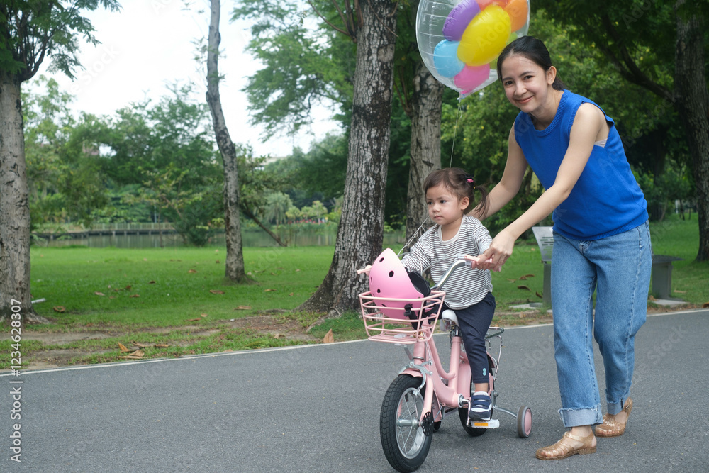 A joyful mother assists her child riding a pink bicycle, surrounded by greenery and colorful balloons in a park setting. A joyful mother assists her child riding a pink bicycle, surrounded by greenery and colorful balloons in a park setting.