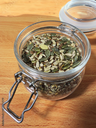 Closeup of mixed edible seeds (sunflower, pumpkin, hemp and linseed) in a nearly full, open glass storage jar, with an airtight lid and metal clasp. 