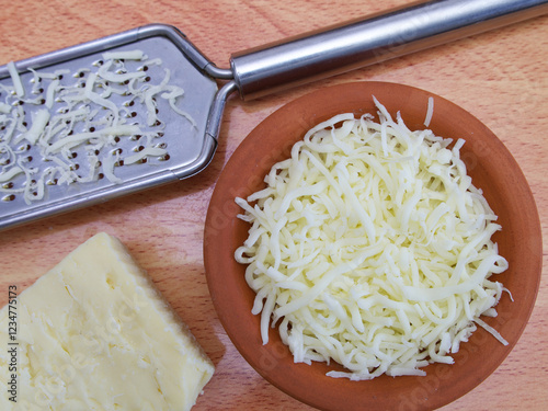 Closeup overhead shot of a steel cheese grater and a piece of cheese, next to a clay dish filled with grated cheese.