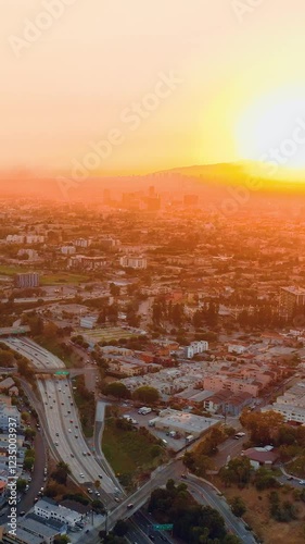Los Angeles cityscape with streets, cars and houses in the rays of a large yellow sun setting over the horizon. Aerial evening view. Vertical video.